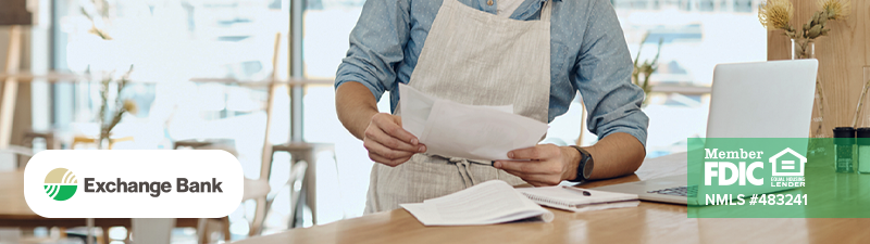 Small business owner wearing an apron looking at paperwork at their desk with laptop in front of them